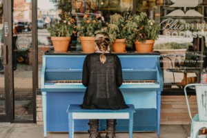 This is a photo of a young woman in a dark long dress with a single braid down her back, sitting at a bright blue painted spinet piano. Her back is to the viewer. She and the piano are outside the glass walls of a storefront, "Bluebird Books" and 4 terracotta planters with flowering plants are arranged in a line on top of the piano she is playing.