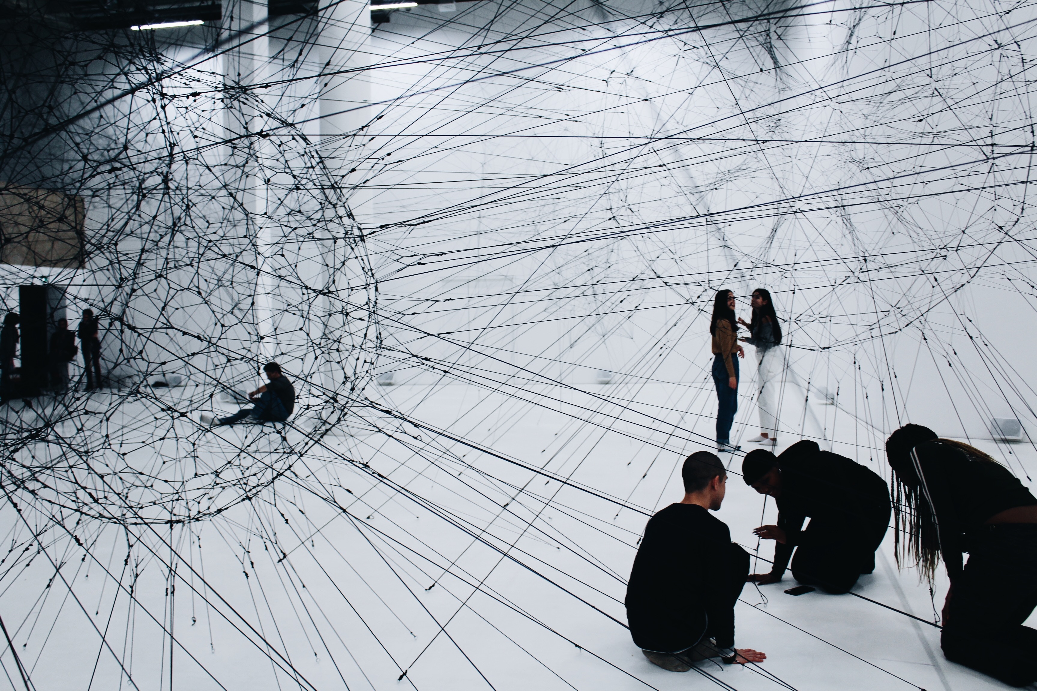 This is a photo of people dressed in all-black clothing in a white room or space. Black thin cords and cables are being connected in networks, and those are connected to two large orbs created by the webs of cords and cables. This photograph was taken by Alina Grubnyak.