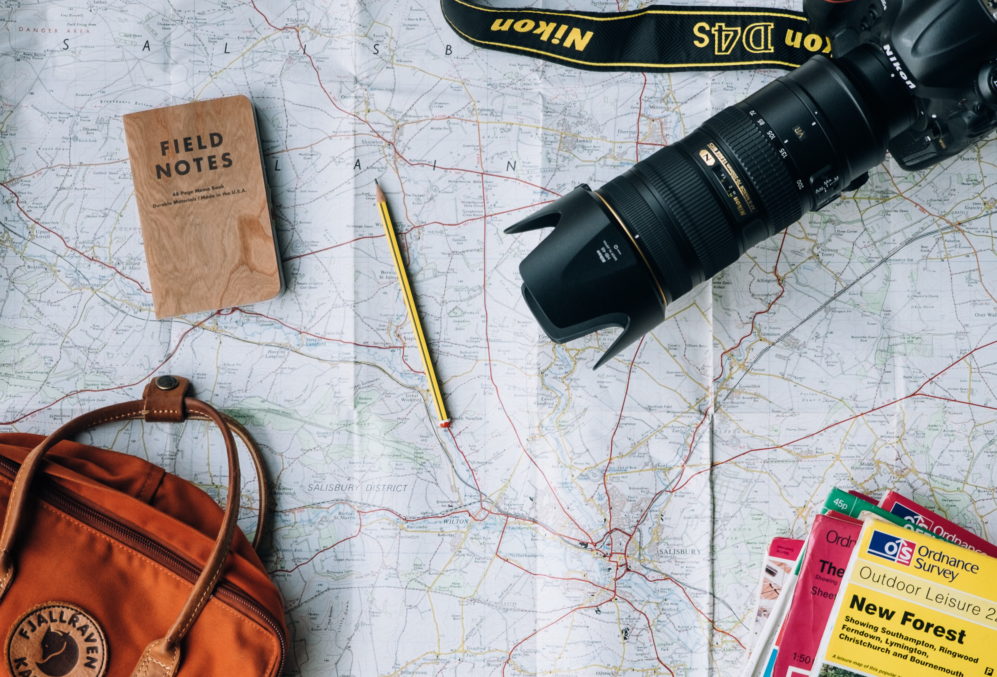 A Nikon camera with a zoom lens, a pencil and a field notebook, a guidebook and a travel bag are strewn across a printed map on a table. This photograph was taken by Annie Spratt.