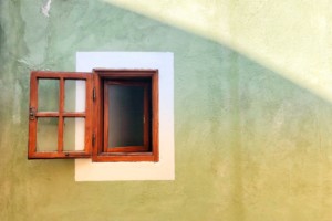 A small square window with a wood frame and wood panes is open. The view is from the outside of a plain concrete building with only the window shown off to the left side. This photograph was taken by Katerina Pavlyuc.