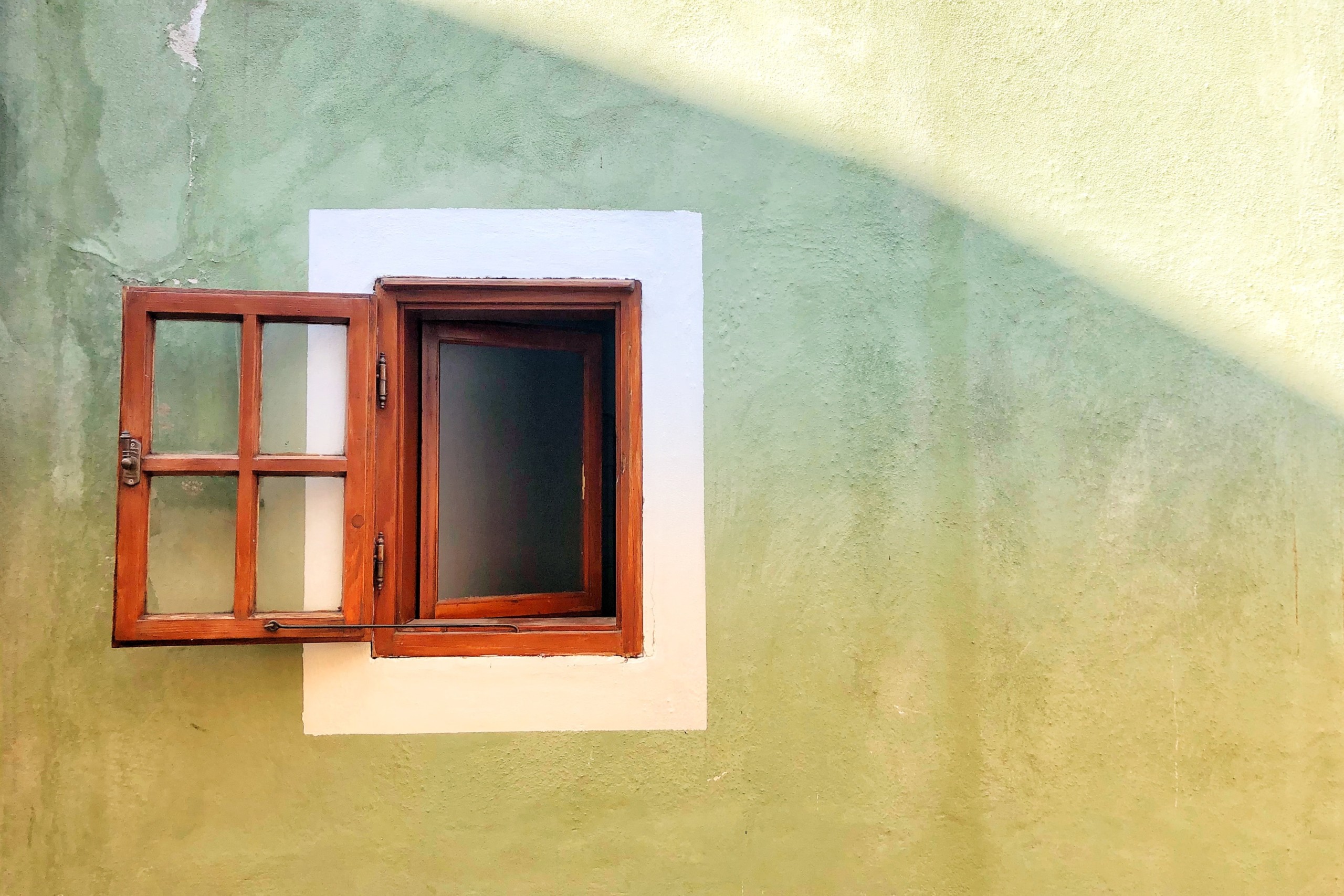 A small square window with a wood frame and wood panes is open. The view is from the outside of a plain concrete building with only the window shown off to the left side. This photograph was taken by Katerina Pavlyuc.